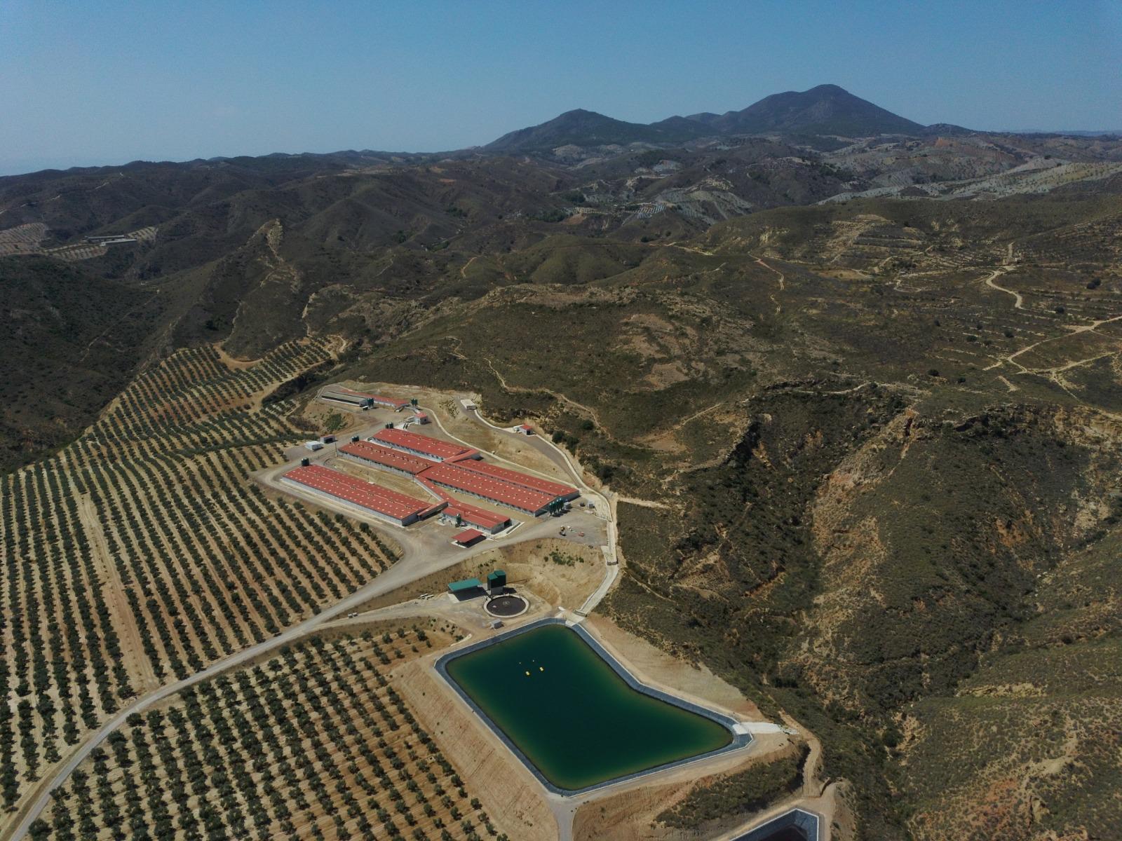 Vista aérea de un complejo agroganadero con varias naves industriales, una balsa de acumulación de agua y campos de cultivo en un entorno montañoso
